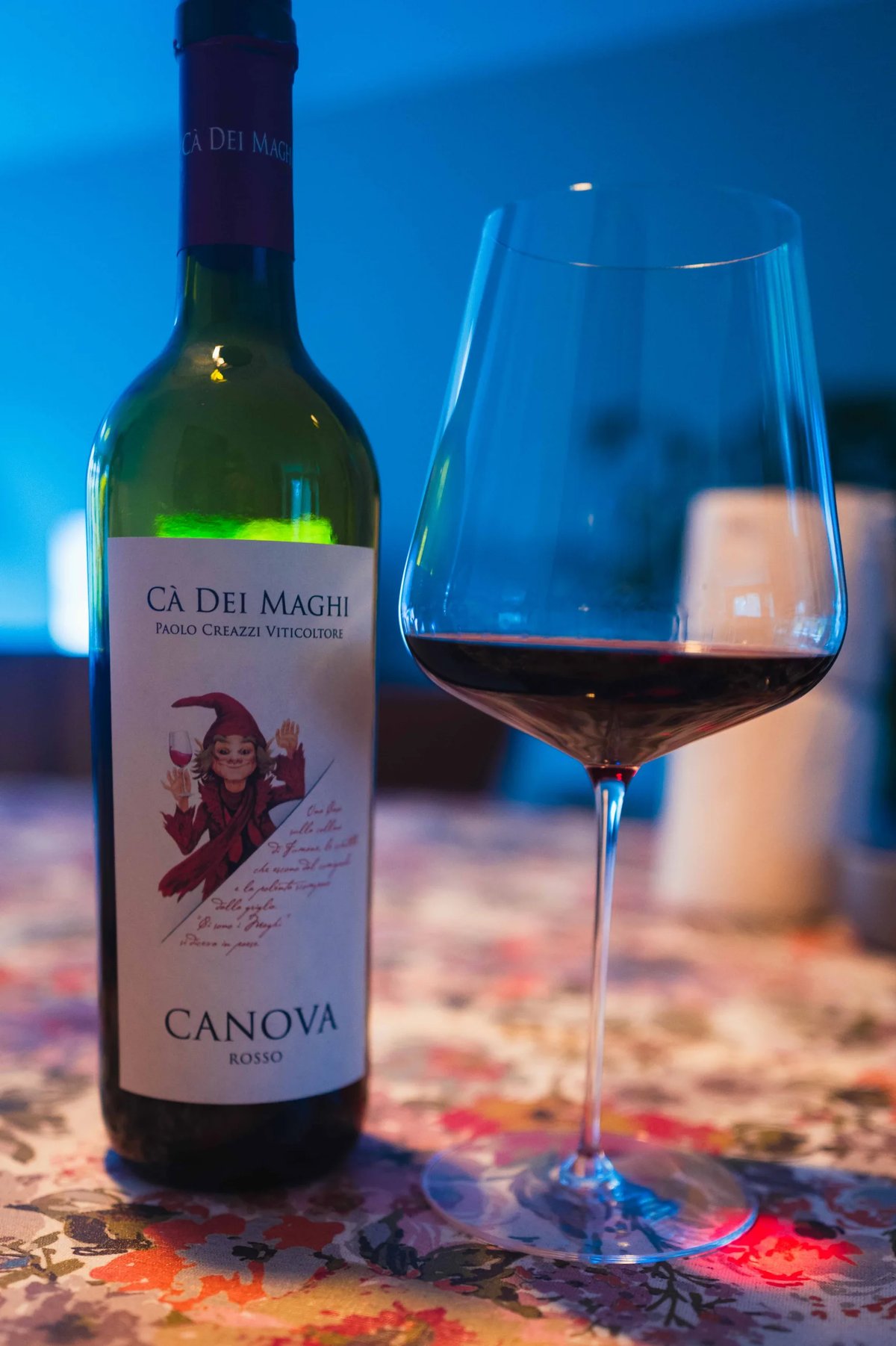 Fles Cà dei Maghi Canova Rosso naast glas rode wijn op bloemenkleed in blauw licht