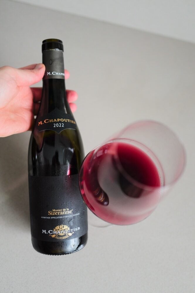 Hand houdt fles M. Chapoutier Sizeranne Hermitage schuin naast liggend glas rode wijn