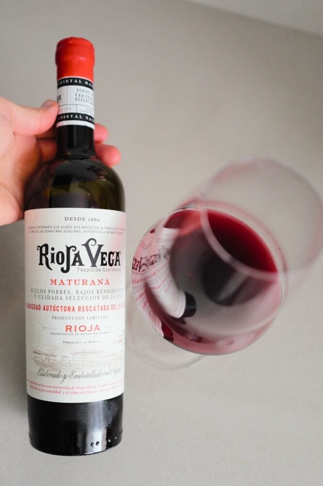 Hand houdt fles Rioja Vega Maturana schuin naast liggend glas rode wijn