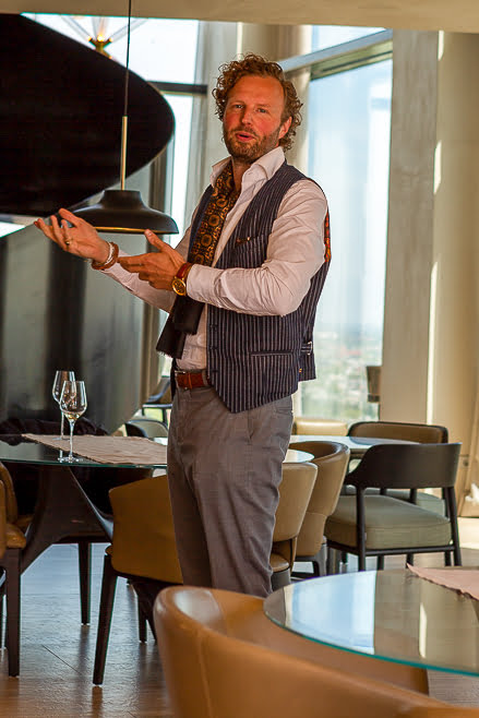 Man in gestreept gilet vertelt staand bij een tafel met witte wijn aan het raam
