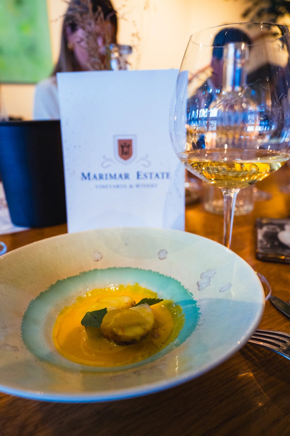 Gerecht in een turquoise bord met Marimar Estate menukaart en een glas goudkleurige wijn