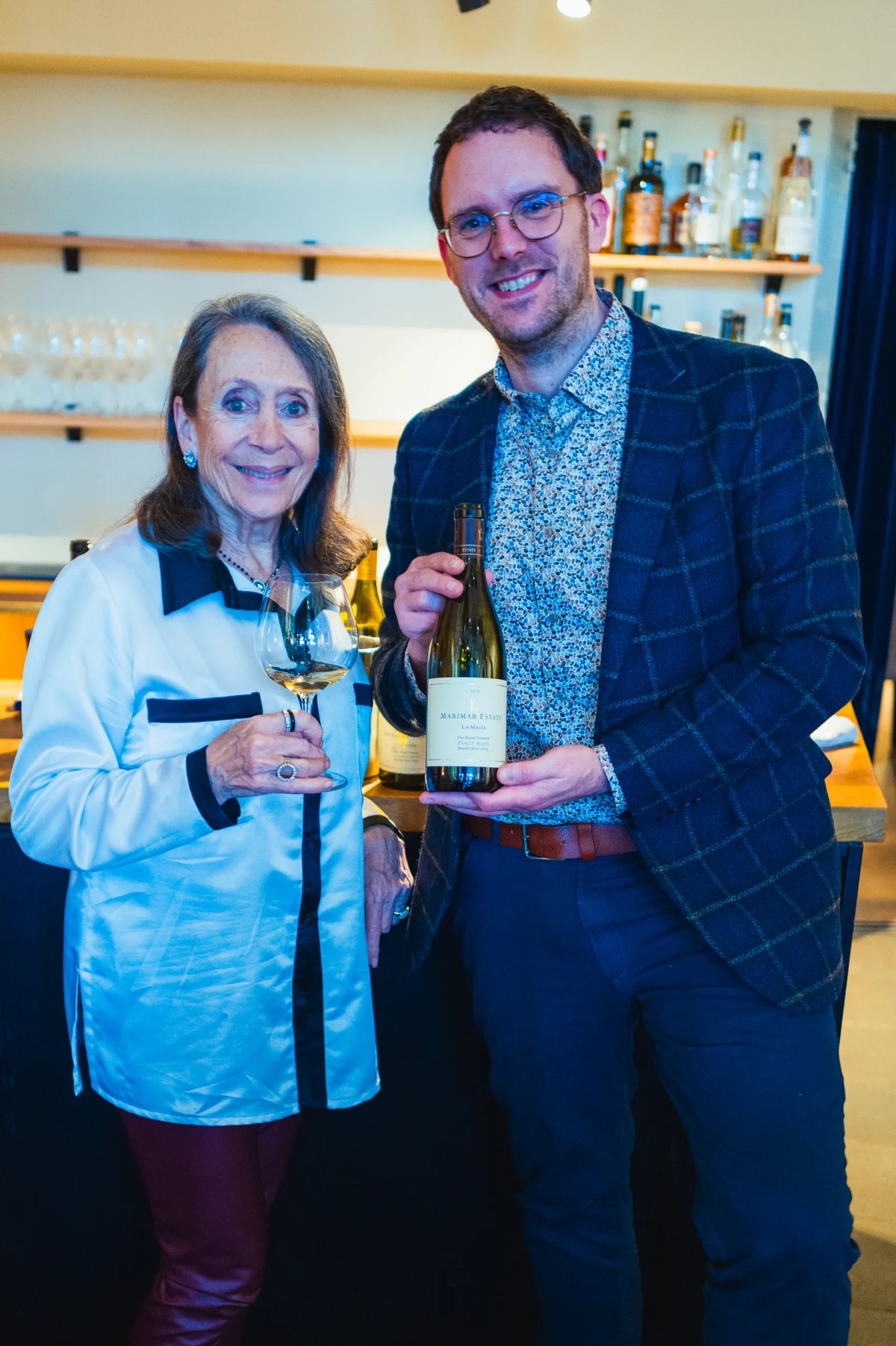 Vrouw in witte jas met een glas en man met bril die een witte wijnfles toont