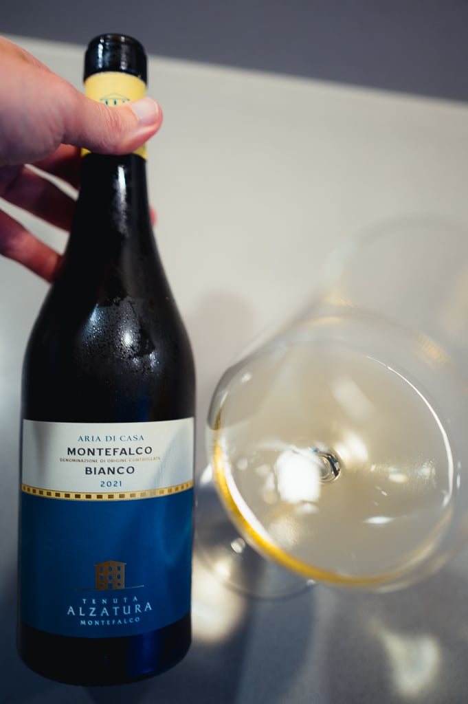 Hand houdt een fles Aria di Casa Montefalco Bianco bij een gekanteld glas witte wijn