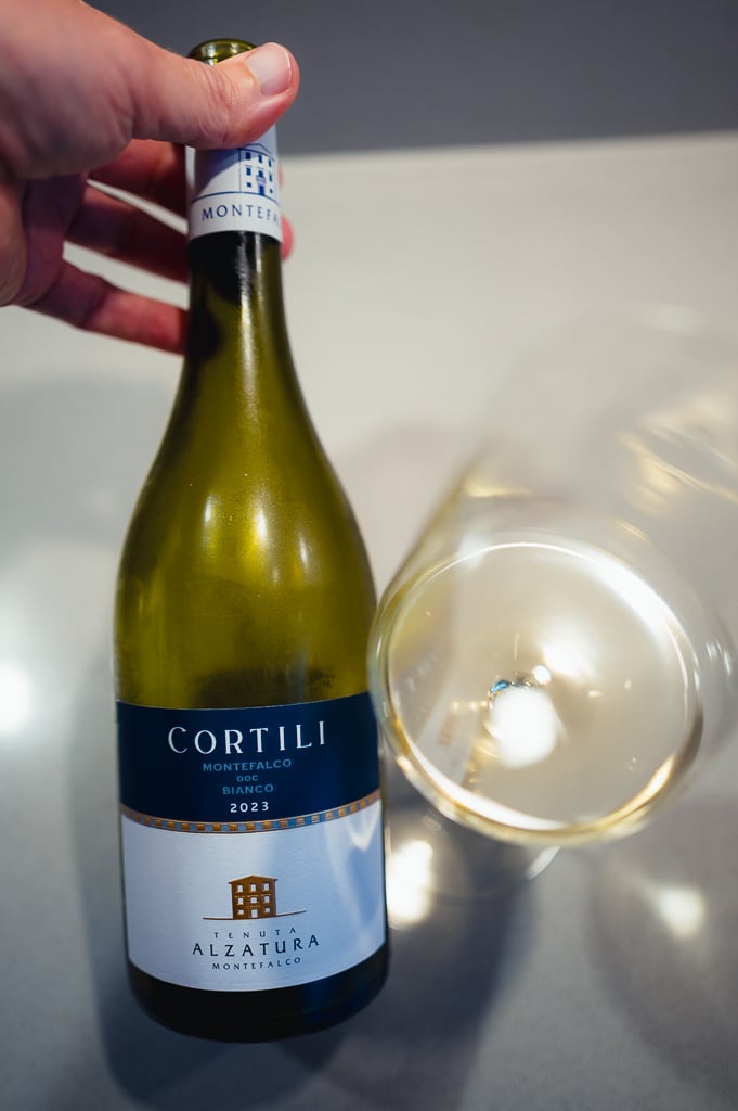 Hand houdt een fles Cortili Montefalco Bianco bij een gekanteld glas witte wijn