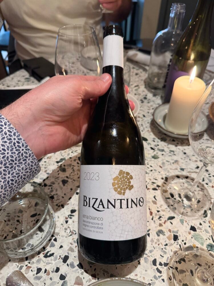 Hand houdt een fles Bizantino Etna Bianco close-up boven een terrazzotafel
