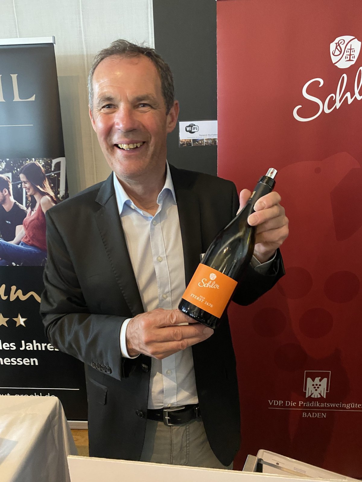 Lachende man in blazer houdt een fles Schlör Fyerst 1476 bij een rode beurswand
