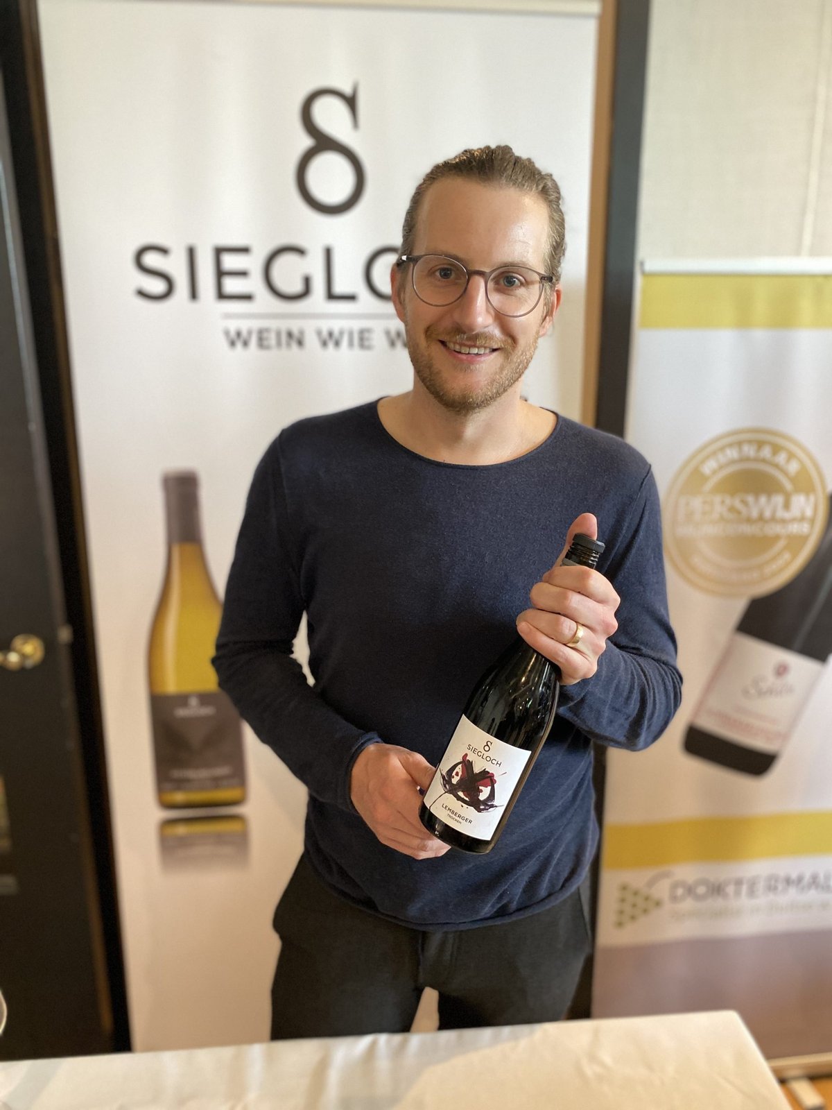Lachende man in blauwe trui houdt een fles Siegloch Lemberger vast