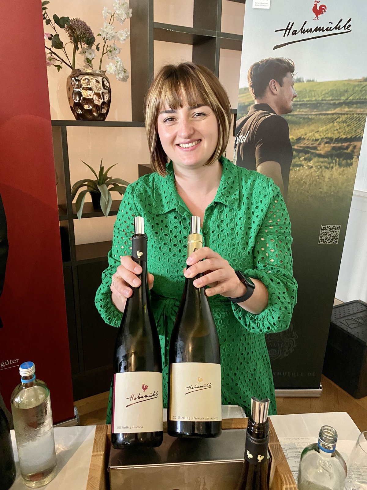 Lachende vrouw in groene jurk houdt twee flessen Hahnmühle Riesling vast