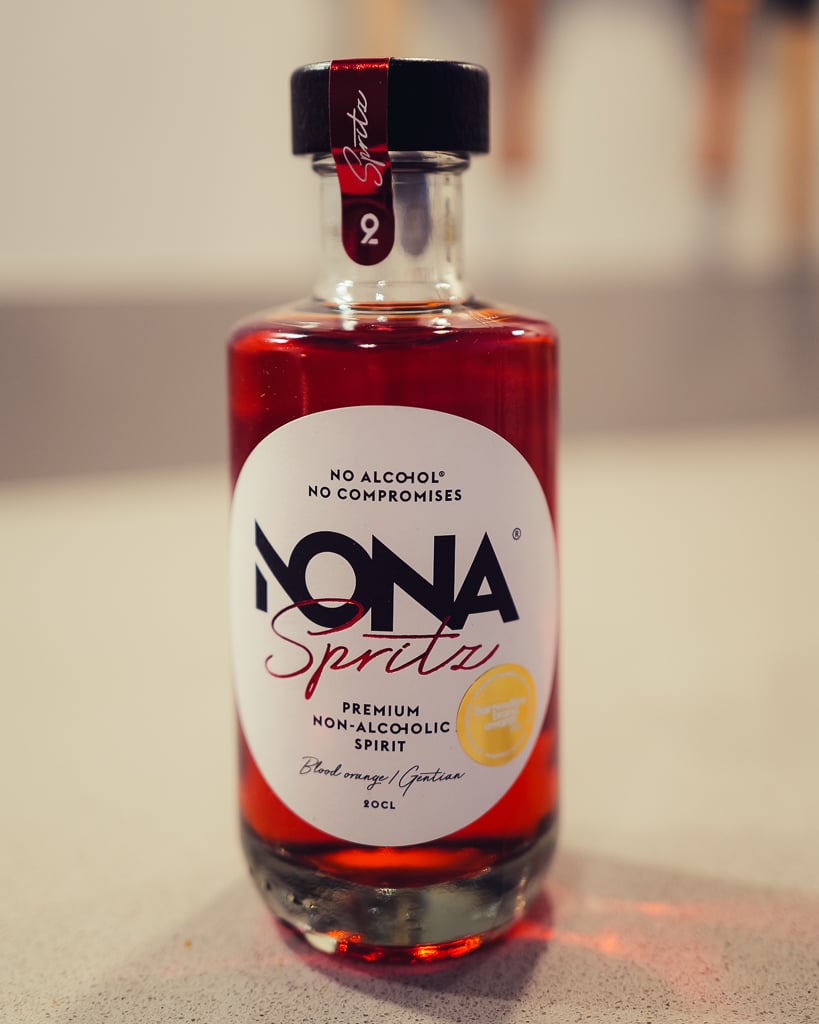 Fles NONA Spritz met dieprode vloeistof naast een glas met bloedsinaasappelkleurige drank en ijs