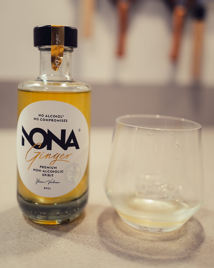 Fles NONA Ginger alcoholvrije spirit met goudgele vloeistof naast een leeg glas op tafel
