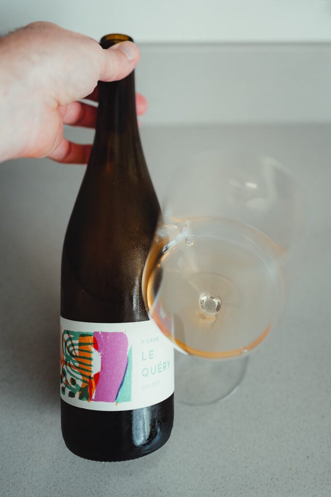 Hand houdt fles Pierre Goiset Le Quéry naast een glas roodoranje natuurwijn