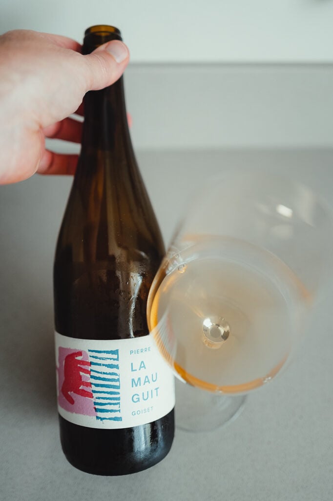 Hand houdt fles Pierre Goiset La Mauguit naast een glas oranjekleurige natuurwijn