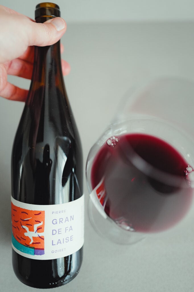 Hand houdt fles Pierre Goiset Gran de Falaise naast een glas helderrode natuurwijn