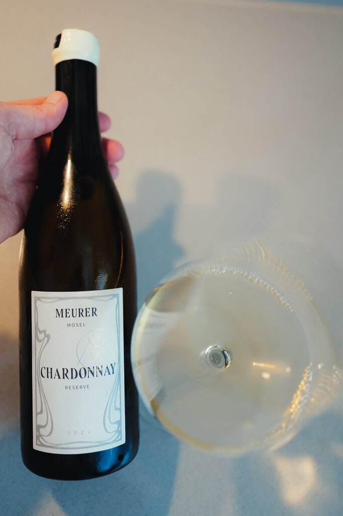 Hand houdt fles Meurer Chardonnay Reserve boven een wijnglas, van bovenaf gezien