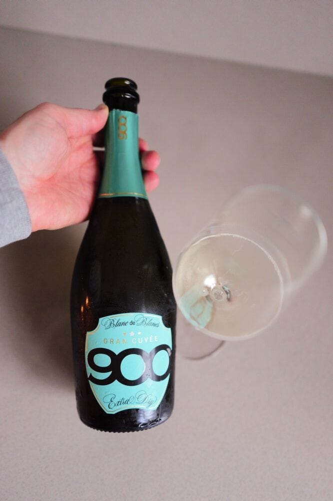 Hand houdt fles 900wine Blanc de Blancs Prosecco met turquoise etiket boven een liggend glas