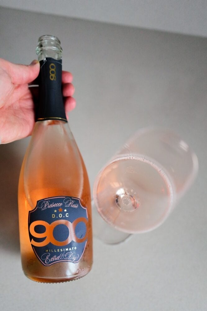 Hand houdt fles 900wine Prosecco Rosé met roze inhoud naast een liggend glas