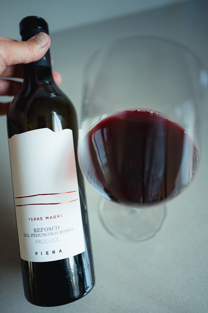 Hand houdt fles Piera Refosco dal Peduncolo Rosso naast een glas dieprode wijn