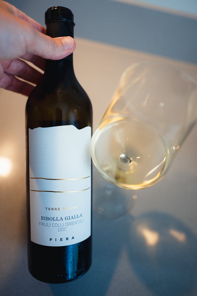 Hand houdt fles Piera Ribolla Gialla naast een glas lichtgele wijn