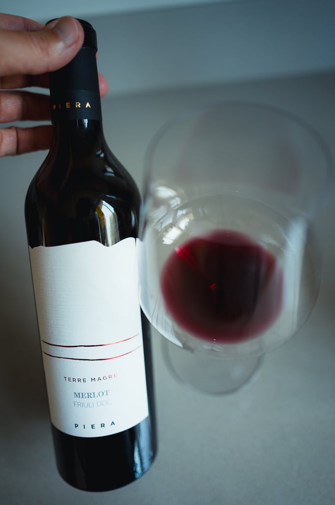 Hand houdt fles Piera Merlot uit Friuli naast een glas dieprode wijn