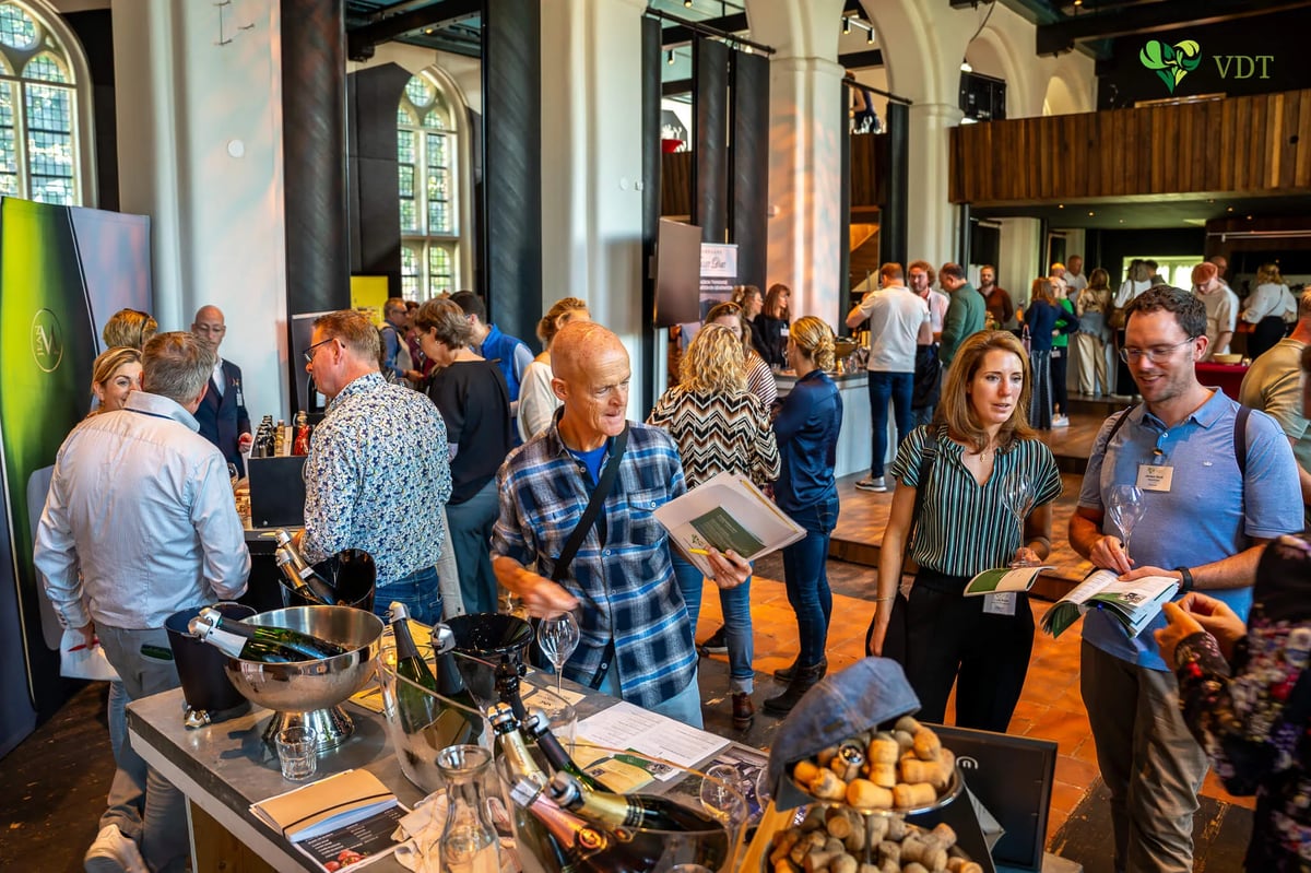 Drukke champagneproeverij in een hoge zaal met bezoekers en flessen in koelers