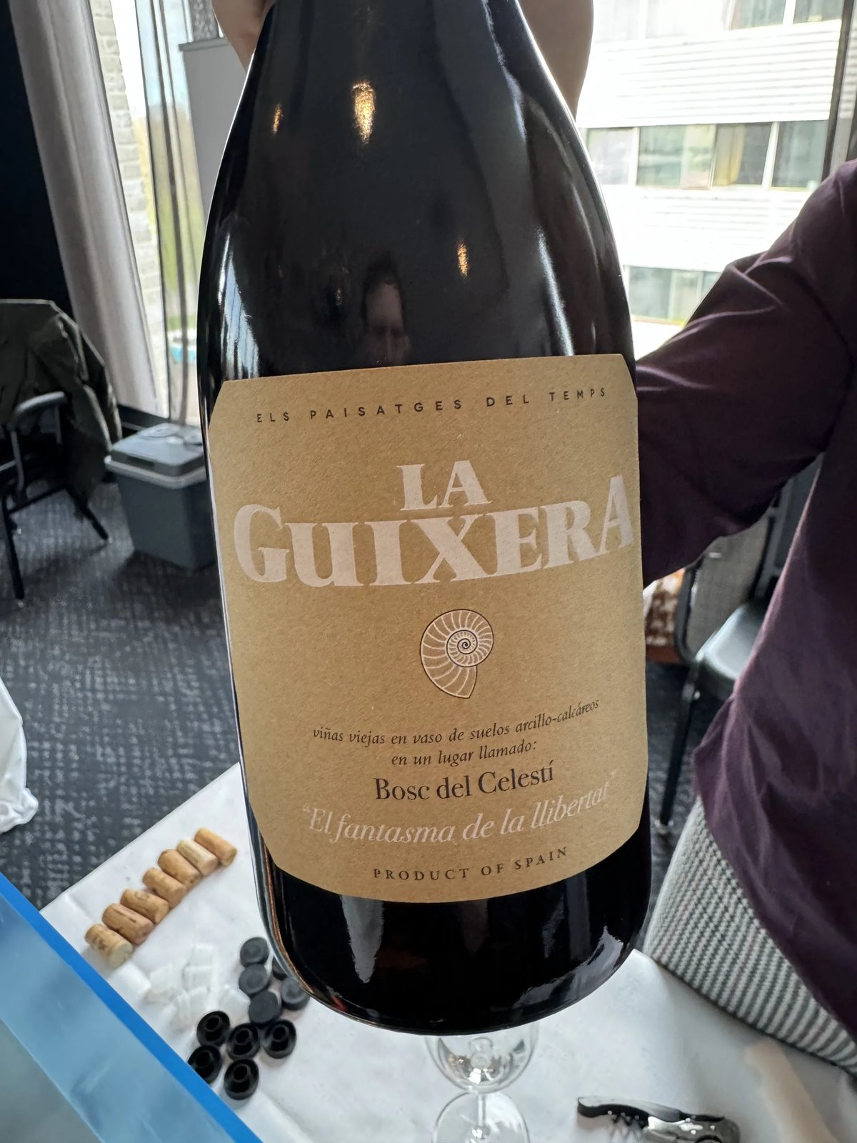 Fles La Guixera met kraftpapier-etiket en schelpmotief, vastgehouden bij een raam