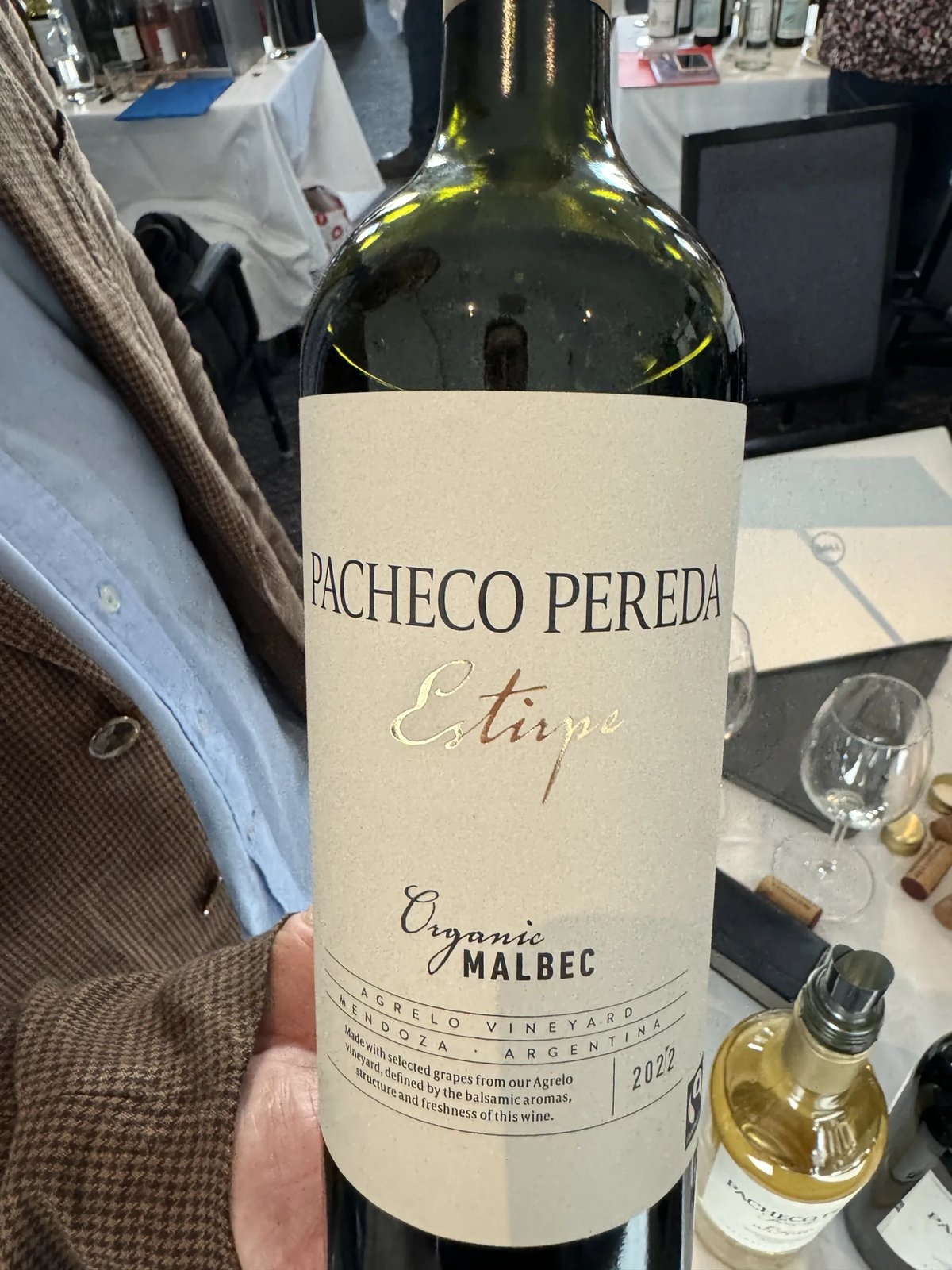 Hand houdt fles Pacheco Pereda Estirpe Organic Malbec 2022 vast bij een proefstand