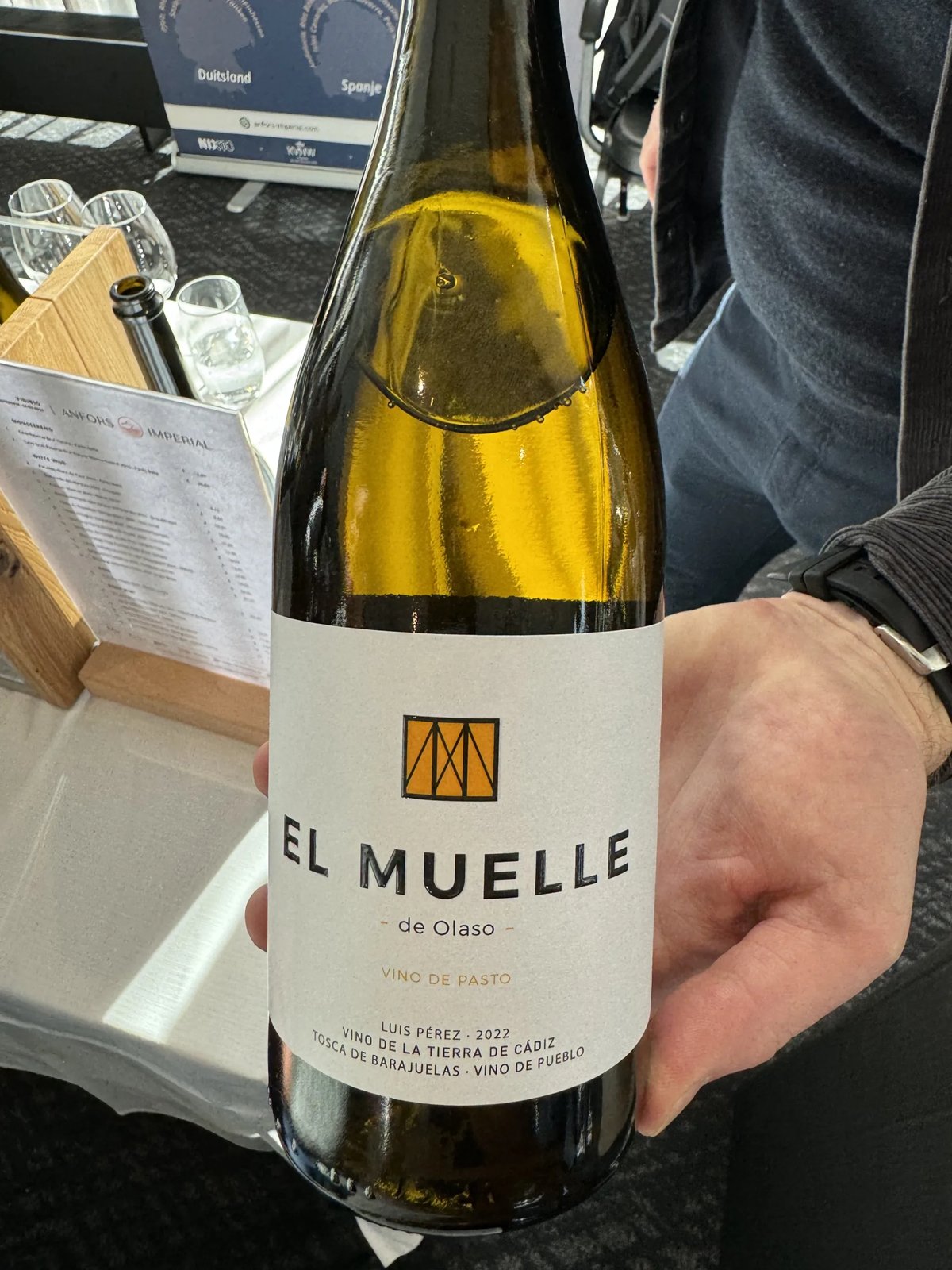 Hand houdt fles El Muelle de Olaso witte wijn vast aan een proeftafel
