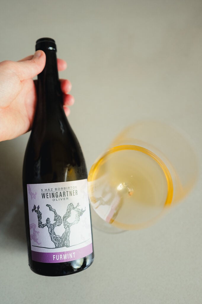 Hand kantelt fles Weingärtner Furmint naast een liggend glas oranje wijn van bovenaf