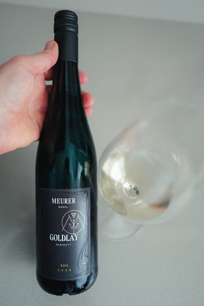 Hand houdt fles Meurer Goldlay Kabinett vast naast een liggend glas witte wijn