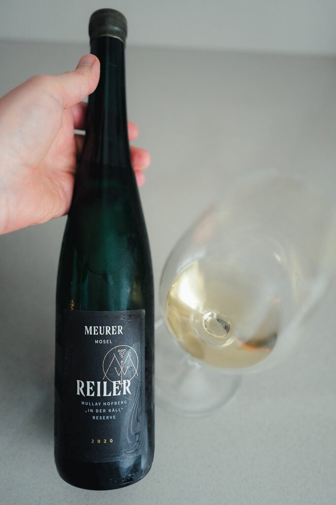 Hand houdt donkergroene fles Meurer Reiler Riesling vast naast een glas witte wijn