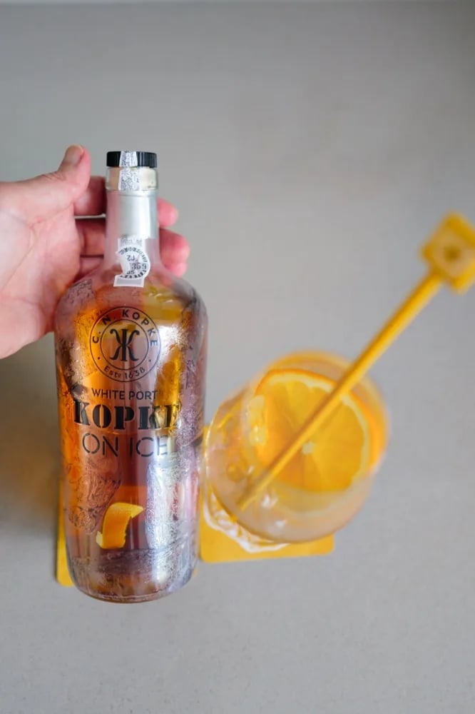 Hand houdt een glas Kopke On Ice met ijs en een schijf sinaasappel naast de fles