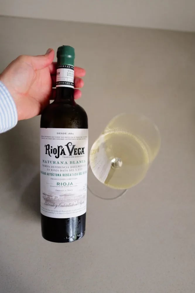 Hand houdt de fles Rioja Vega Maturana Blanca 2025 vast naast een gekanteld glas witte wijn