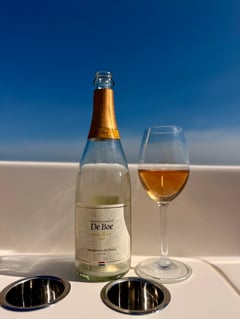 Fles Wijndomein De Boe Rosé Brut 2023 naast een glas koperkleurige mousserende wijn op een boot, met het water van de Oosterschelde erachter
