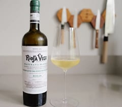 Fles Rioja Vega Maturana Blanca 2025 met een glas witte wijn op een keukenblad, Japanse messen aan de wand