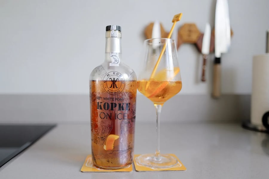 Fles Kopke On Ice witte port naast een wijnglas met drie ijsblokjes en een sinaasappelschil op een keukenblad