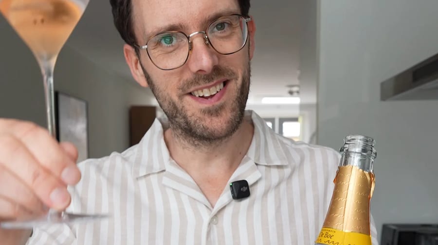 Fles Wijndomein De Boe Rosé Brut 2024 naast een glas koperkleurige mousserende wijn, geheven voor een proost